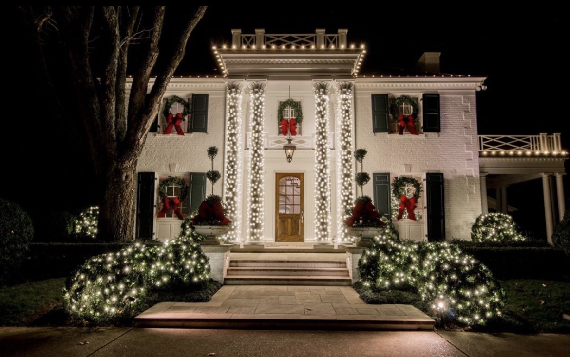 White columned home in Short Hills NJ with every column wrapped in warm white lights, red-bow wreaths in the windows and lit boxwoods along the front walk